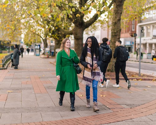 A couple of women walking down a leafy pavement, talking and laughing