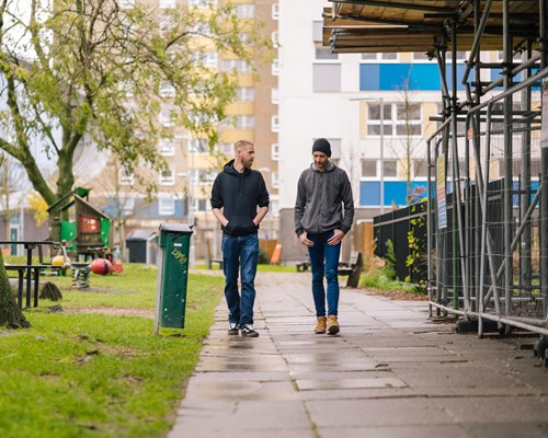 Two men walking and talking on a path that goes through a green area in what is an otherwise built up urban area