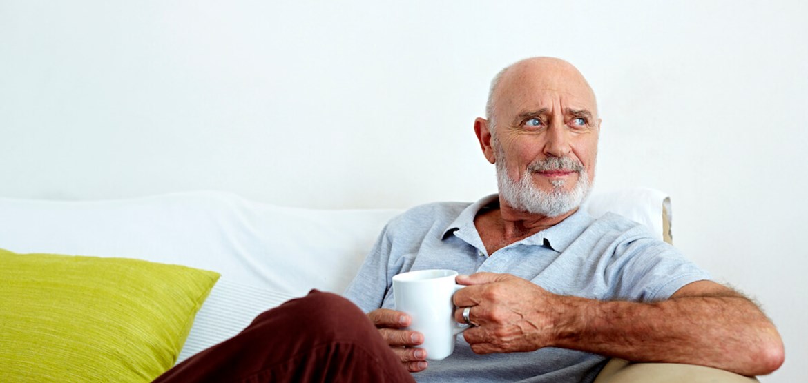 A man sitting on a sofa with his leg crossed, holding a cup while looking off to the left