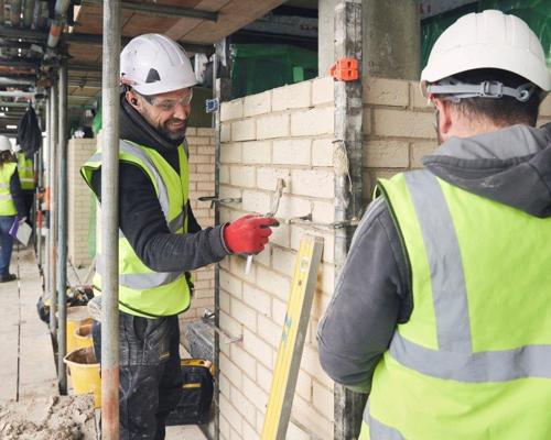 Two men in high vis jackets and hard hats stood in a construction area with a wall between them. One of the men is holding a trowel with cement