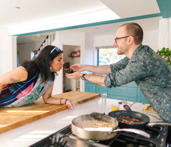 A woman leaning over a counter top in a kitchen to taste some food that a man is cooking