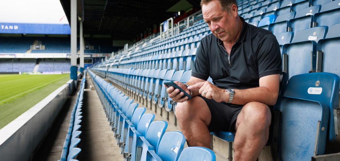 Footballer David Seaman sat on the bleachers in a football stadium looking at his phone