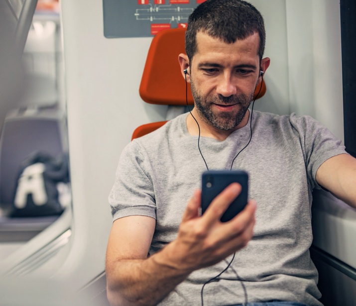 A man sat on a train with headphones in his ears holding his phone in his right hand looking at it