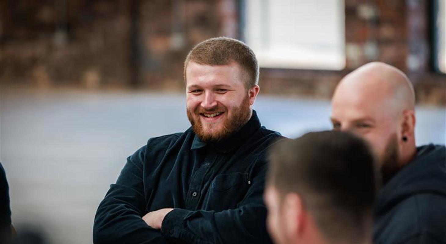 A man sitting with his arms crossed, smiling while listening to other men talk