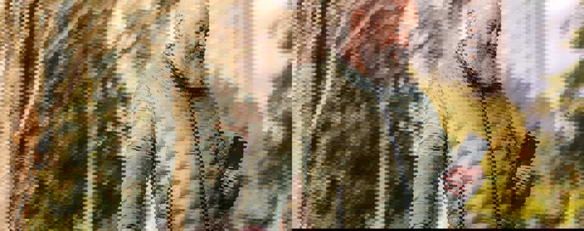 A middle-aged man in a green jacket looking at his phone with a blurred background with trees