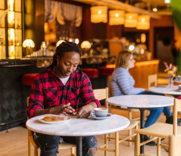 Person sat at table in cafe with coffee and a pastry, looking at their phone.