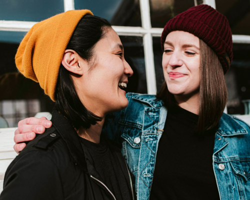 Two women, wearing beanies, looking at each other and smiling