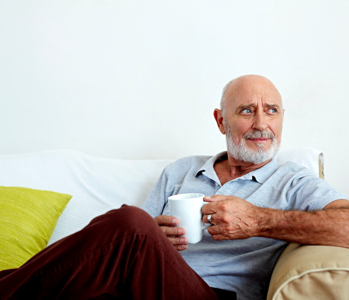 A man sitting on a sofa with his leg crossed, holding a cup while looking off to the left