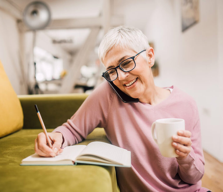 Woman sitting on the floor next to a sofa, holding her phone to her face with her shoulder. She is making notes in a notebook that rests on the sofa