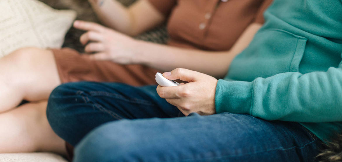 Two people sitting on a sofa, heads are out of shot. One is taking glasses off their face while the other is searching on their phone