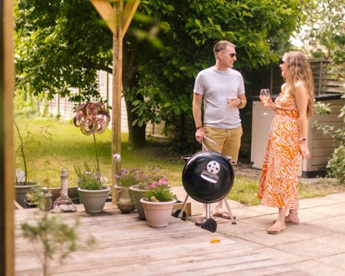 A couple in their garden, stood by a barbecue, with wine glasses in had.