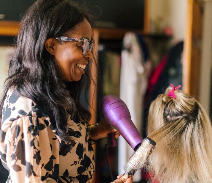 Person blow-drying friends hair in bedroom.