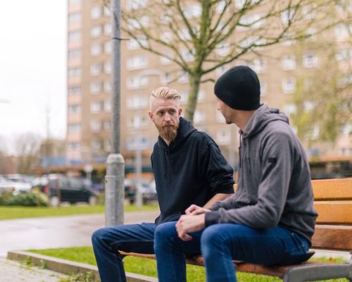 Two people chatting on a bench in front of a block of flats.